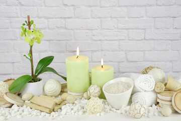 spa still life of soft green orchid flower plant with burning candles surrounded in natural dried elements, white towels, bath salts against a  white brick background.