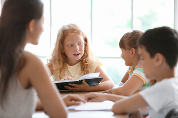 Little redhead girl reading book during literature lesson in classroom
