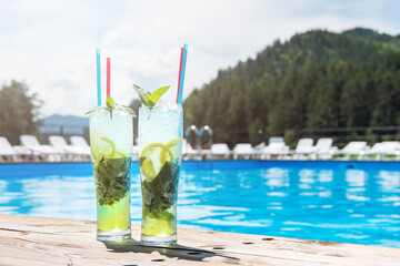 Two Mojito cocktail with lime and mint in highball glass at the swiming pool background on summer mountain background. Concept of summer relaxing