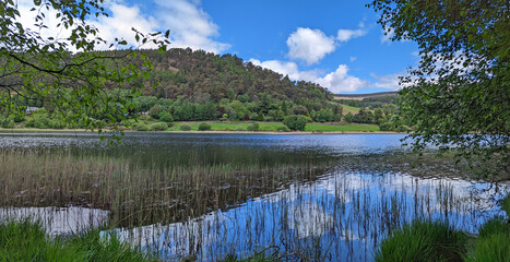 View across the water along Lower Lake in the Glendalough Valley in the Wicklow Mountains National Park in Ireland with a cloudy blue sky, green forest and sky reflecting water