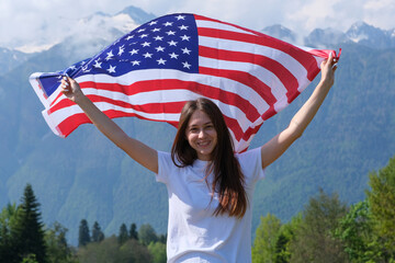 woman holding american flag 
A young smiling woman, a brunette in a white t-shirt, holds an American flag in her hands held high. Outside the house, in the mountains, against the sky.