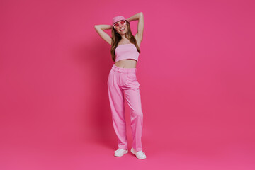 Full length of attractive young woman in trendy clothing standing against pink background