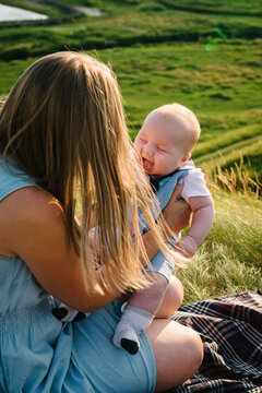 Funny baby. Mother with happy son boy sitting on a blanket at picnic in nature. Child fun on the green grass in the field at sunset. Kid is playing and smiling in the mountains. Close up.