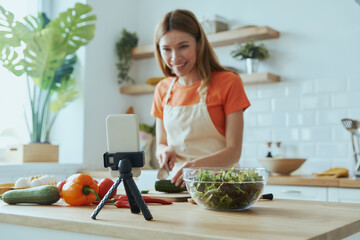 Beautiful young woman cooking and vlogging while standing at the domestic kitchen