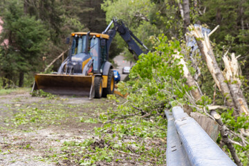 Selective focus view on a rural highway in the aftermath of powerful storm, blurry excavator is seen at work in background clearing fallen trees.