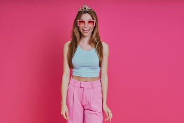 Happy young woman in funky crown looking at camera while standing against pink background