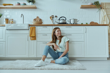 Happy young woman holding mobile phone while sitting on the floor in the kitchen