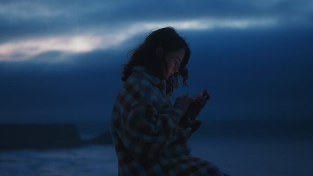 Soft cinematic focus on dreamy millennial girl sits on ocean shore on dark windy Porto night, looks at smartphone, texting, chatting with boyfriend, girlfriend with blue clouds and waves on background