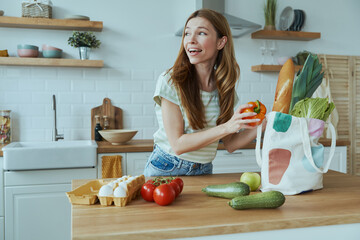 Beautiful young woman unpacking the bag with healthy food while standing at the domestic kitchen - obrazy, fototapety, plakaty