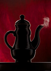 Antique black ceramic coffee pot on a red background, close-up. Kettle with puffs of steam from the spout. boiling. Teapot with boiling water