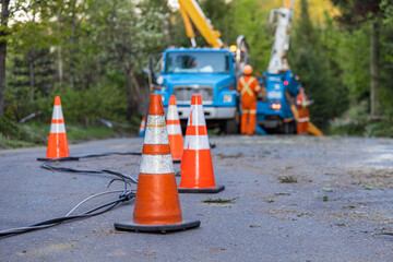 Selective focus view of high viz road cones warning traffic of fallen overhead power line after storm. Blurry contractors work to restore electricity. - obrazy, fototapety, plakaty