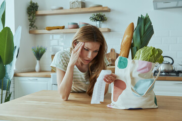 Depressed young woman checking receipts while standing at the domestic kitchen