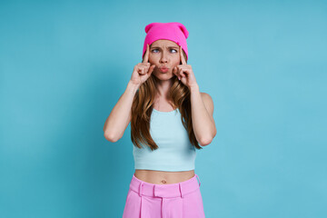 Playful young woman in pink hat grimacing while standing against blue background