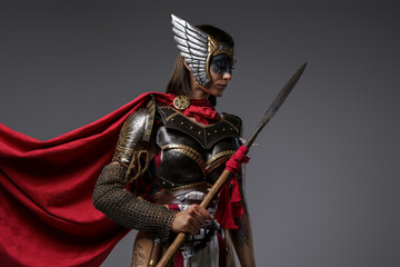 Obraz premium Shot of female barbarian holding spear dressed in steel armor with helmet against grey background.