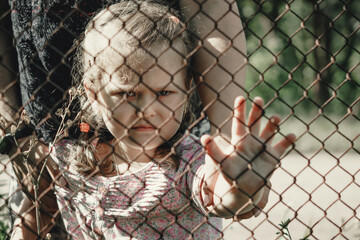 A little girl with a sad look behind a metal fence. The problem of forced deportation of Ukrainian...