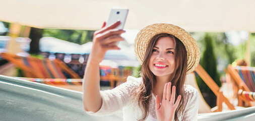 Young woman tourist traveler smiling, waving hand at the phone screen, sitting in a hammock on the...