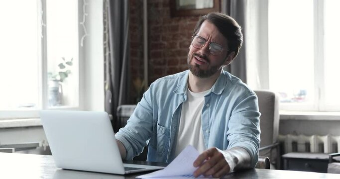 Annoyed millennial man in glasses manage financial documents at home office read reports feel angry upset surprised with bad news failing sales. Distressed young businessman receive tax penalty paper