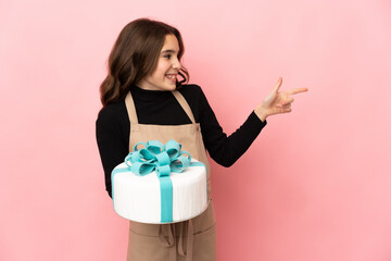 Little Pastry chef holding a big cake isolated on pink background pointing finger to the side and presenting a product