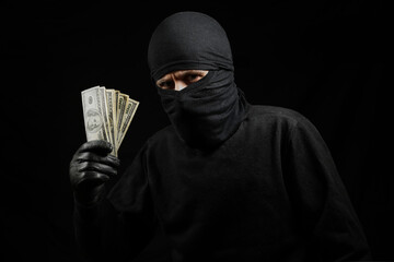 thief in a black mask with dollars in hands on a black background. robber in a black balaclava with...