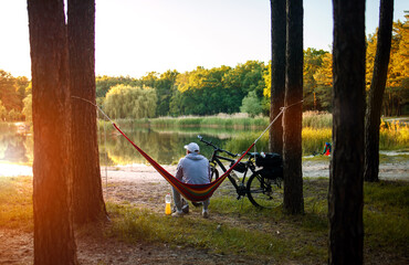 Man travels on bicycle, relaxing in red hammock, rest in forest near lake. Cyclist in hammock at...
