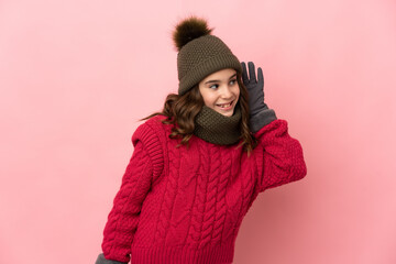 Little girl with winter hat isolated on pink background listening to something by putting hand on the ear