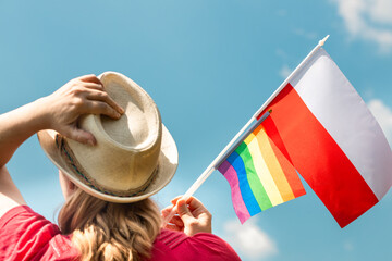 A young woman supporting sexual minorities in Poland proudly holds LGBT and Polish flags in her hands, The other hand holds up a straw hat, against the sky, Concept of the fight for minority rights