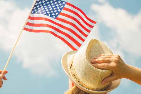 Young Woman Proudly Holds The USA Flag Above Her Head While Holding Up Her Hat With Her Other Hand, Concept Of National Holidays And Patriotic Events