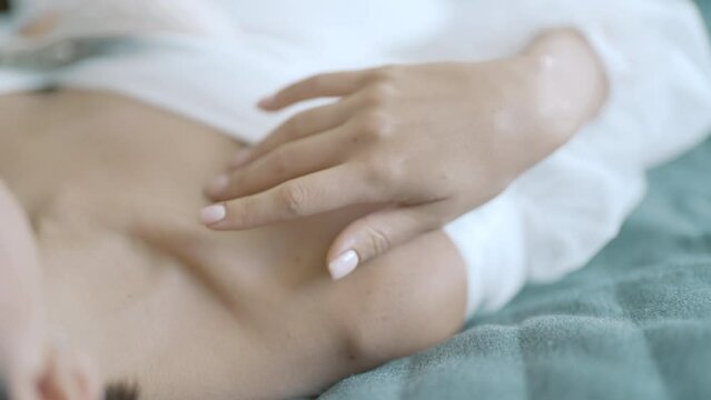 Close up of a young woman touching gently her collarbones while lying on bed in white dress. Action. Tender female relaxing on bed.