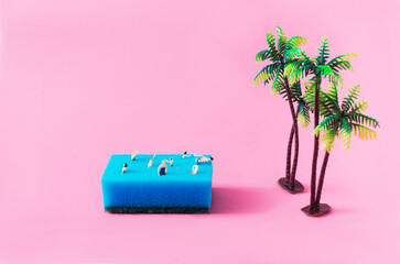 A blue sponge with mini people swimming on it, next to plastic palm trees, against pink background....