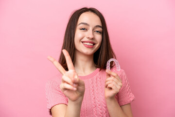 Young Ukrainian woman holding invisible braces isolated on pink background smiling and showing victory sign