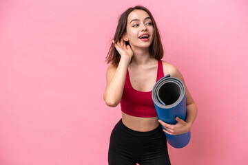 Young sport Ukrainian woman going to yoga classes while holding a mat isolated on pink background listening to something by putting hand on the ear