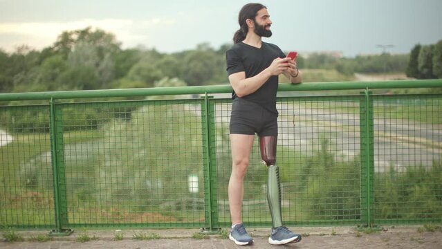 long black hair caucasian bearded man disabled artificial leg smiling and leaning against railing bridge holding mobile phone texting message after workout