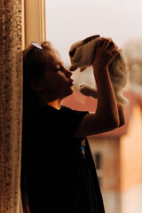 A little girl sits stands on a windowsill in a black T-shirt and sunglasses with her long hair loose at a golden sunset with a rabbit in her hands