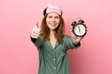 Young English woman in pajamas isolated on pink background in pajamas and holding clock with thumb...