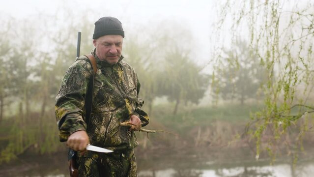 A hunter with a sharp knife sharpens a stick near a misty autumn lake
