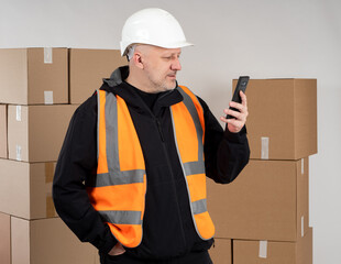 Man with phones. Business man stands in front of boxes. Business owner concept. Guy in orange vest. Business owner uses smartphone. Parcel company owner. Adult human use mobile applications