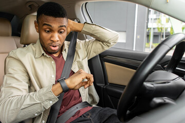 African American guy looking at wrist watch with nervous expression in car
