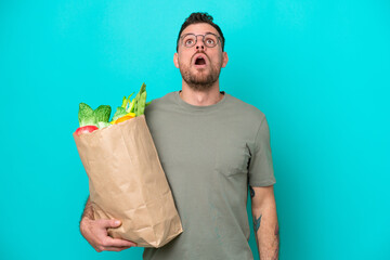 Young Brazilian man holding a grocery shopping bag isolated on blue background looking up and with...