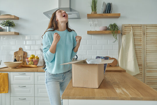 Excited young woman unpacking box and gesturing while standing at the domestic kitchen