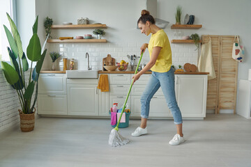 Full length of beautiful young woman cleaning floor with mop while standing at the kitchen