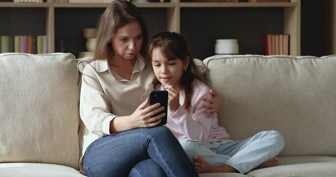 Young generation use modern wireless technology, new mobile application usage concept. Pretty mom her daughter sit on sofa with smartphone, have fun use cool free app spend leisure at home on internet