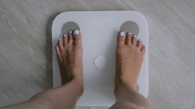 Female bare feet stepping on white digital floor scales - woman weighing herself at home: close up, top view. Measuring weight, control, wellness and diet concept