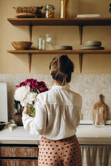 Fototapeta na wymiar a young woman stands with her back with a bouquet of red and white peonies in her hands in the kitchen. a girl in trousers and a white shirt stands with a vase of flowers, view from the back.