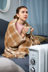 Young adult woman covered in blanket drinking hot tea sitting on sofa near electric heater in cold...