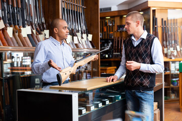 Fototapeta na wymiar Latin american man owner of gun store consulting customer about modern sporting rifle before purchase