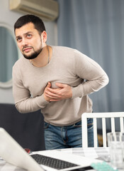 Young man touching his chest with hands while having heart pain and feeling unwell at home,...