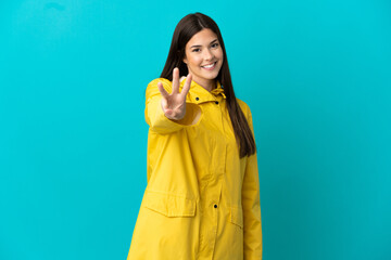 Teenager Brazilian girl wearing a rainproof coat over isolated blue background happy and counting three with fingers