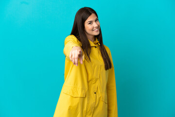 Teenager Brazilian girl wearing a rainproof coat over isolated blue background points finger at you with a confident expression