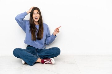 Young woman sitting on the floor surprised and pointing finger to the side