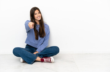 Young woman sitting on the floor points finger at you with a confident expression